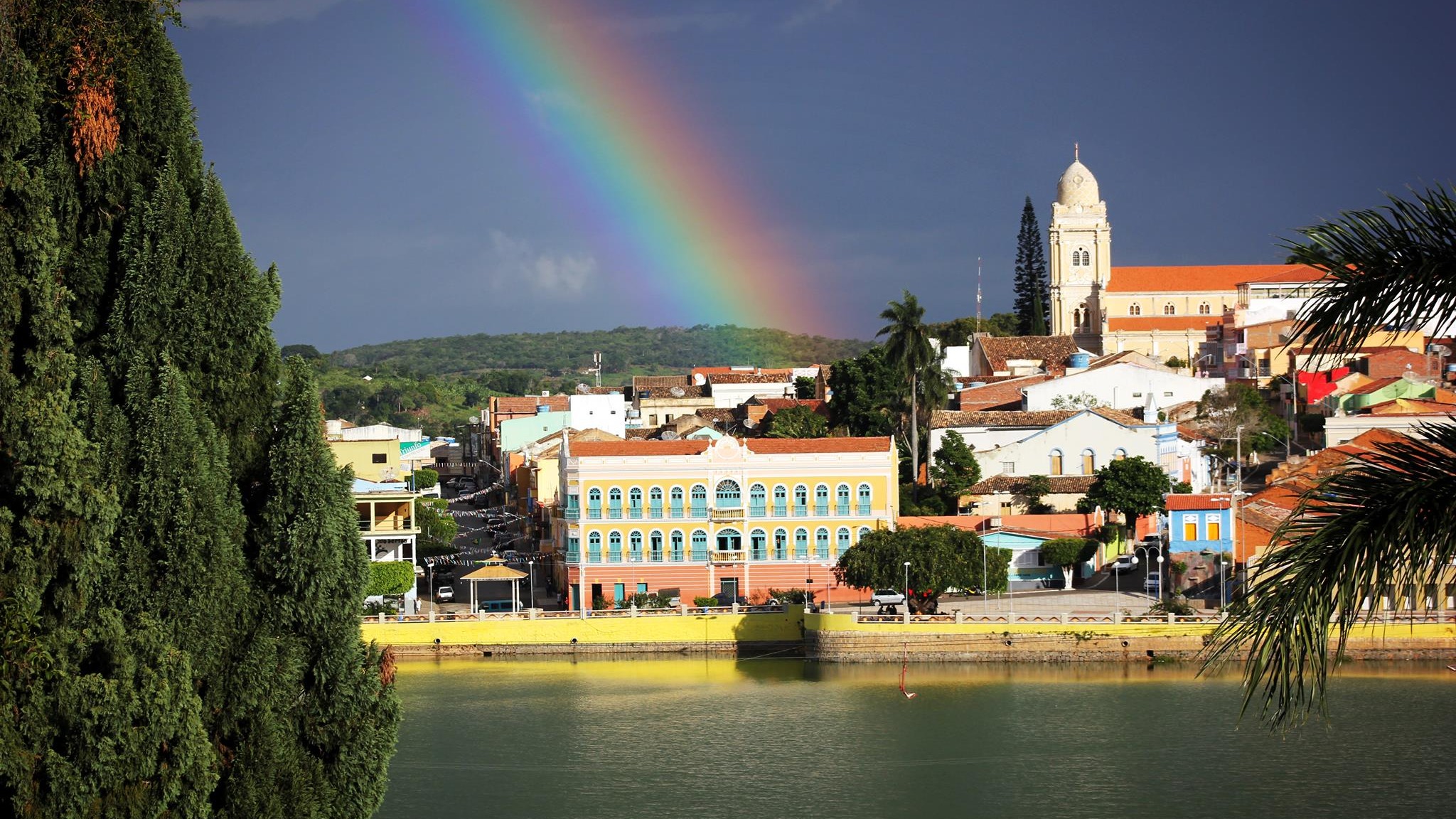 Triunfo, o oásis do Sertão pernambucano, revela sua beleza com o colorido da arquitetura histórica e o Lago João Barbosa emoldurados por um arco-íris. Vista de Triunfo com lago, edifícios coloridos, igreja e um arco-íris vívido no céu.