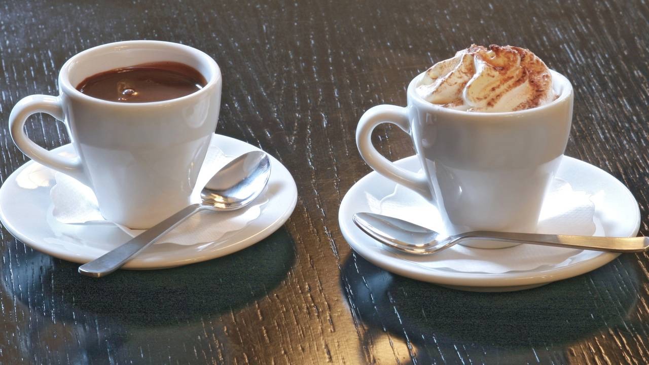 Aprecie uma versão mais leve de chocolate quente para se aquecer nos dias frios sem descuidar da alimentação. Duas xícaras brancas de chocolate quente com colheres em pires sobre mesa de madeira escura