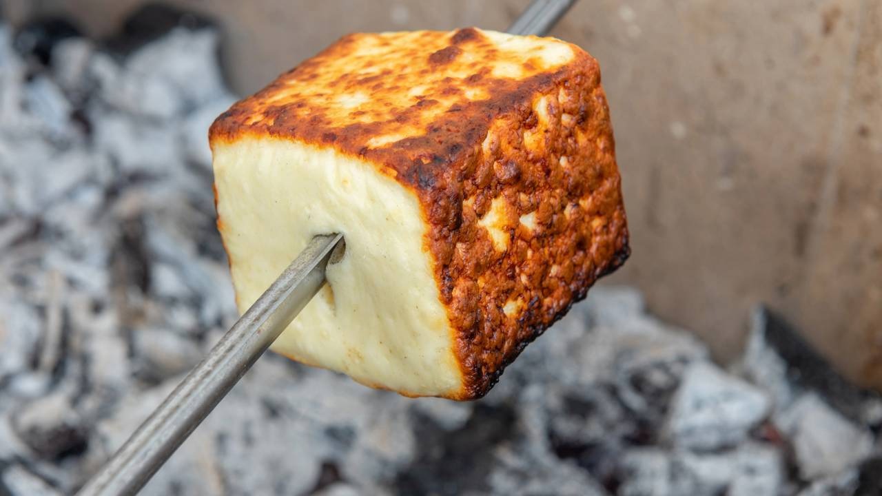Queijo coalho, peça-chave para petiscos saborosos e rápidos, como o enroladinho com bacon na air fryer ideal para a Copa. Bloco de queijo coalho grelhado em espeto de metal, dourado, sobre brasas de carvão.