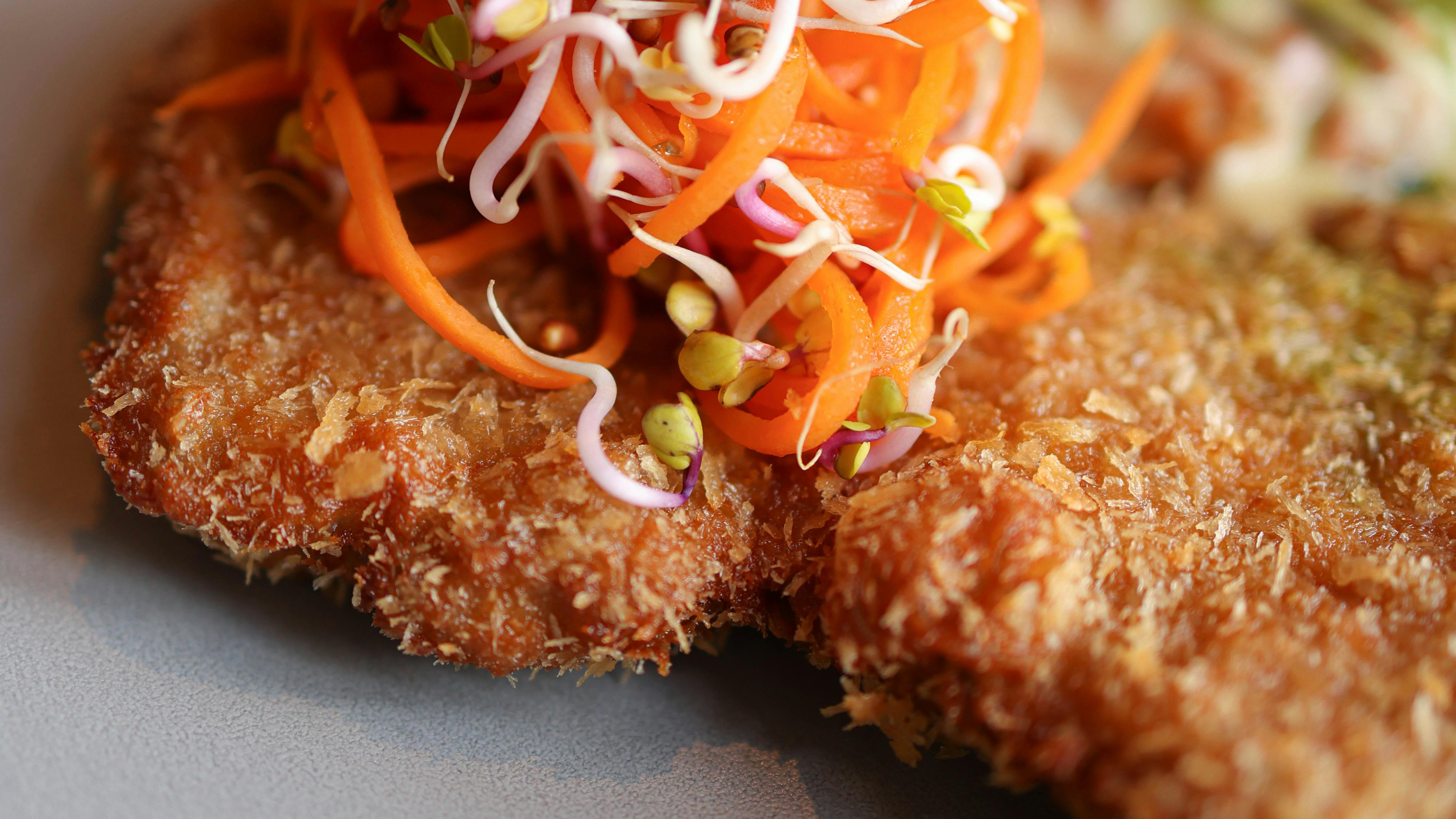 Close-up de tonkatsu empanado e frito, coberto com cenouras julienne e brotos variados.