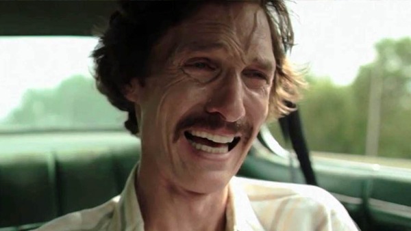 Close-up do rosto de Matthew McConaughey, com cabelo castanho ondulado, bigode e expressão de riso com a boca aberta, revelando os dentes. Há rugas marcadas ao redor dos olhos e na testa. O fundo está borrado, com tons verdes que sugerem o interior de um veículo.