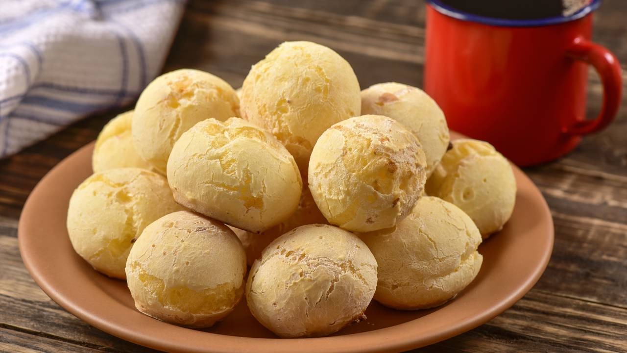 A receita de pão de queijo de ricota e chia é uma das opções leves para o inverno. Bolinhas de pão de queijo em prato marrom com caneca vermelha em mesa de madeira