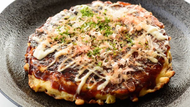 Okonomiyaki redondo com molho escuro, maionese, flocos de peixe seco e alga em pó.