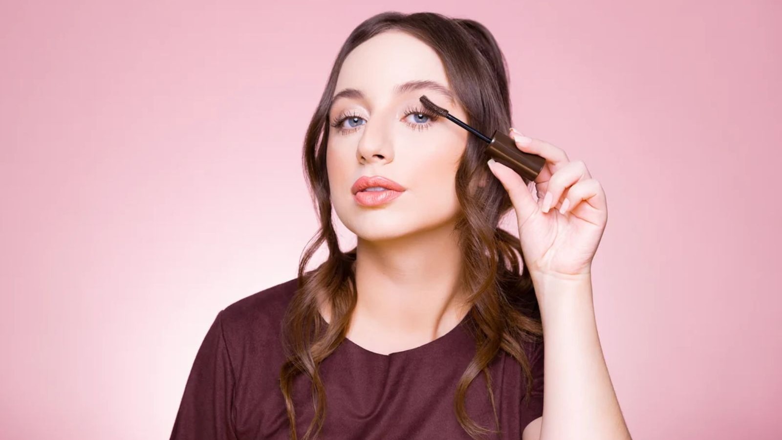 Mulher com cabelo castanho e olhos azuis aplica m&aacute;scara de c&iacute;lios escura em fundo rosa