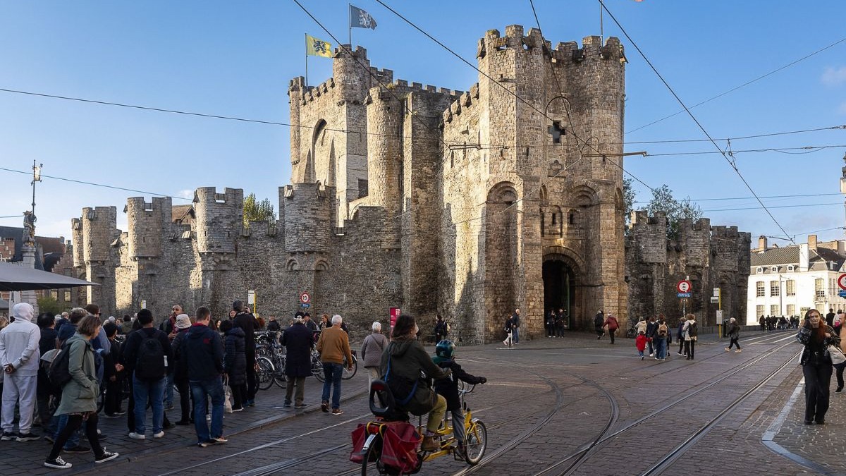 Castelo Gravensteen de pedra, com pessoas, bonde e bandeiras em rua de paralelep&iacute;pedos de Gent.