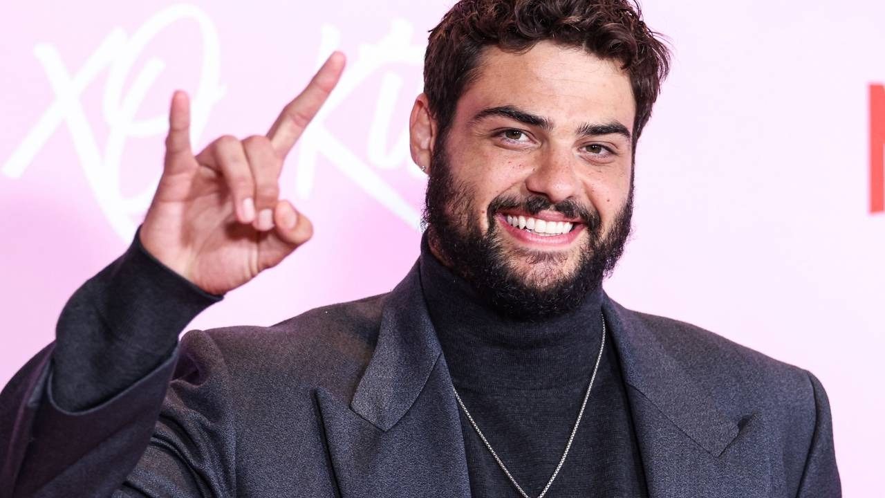 Noah Centineo, galã de "Para Todos os Garotos que Já Amei", que se transformou para seu novo papel como Rambo. Noah Centineo sorrindo, com barba, blazer escuro e gola alta, fazendo sinal com a mão.