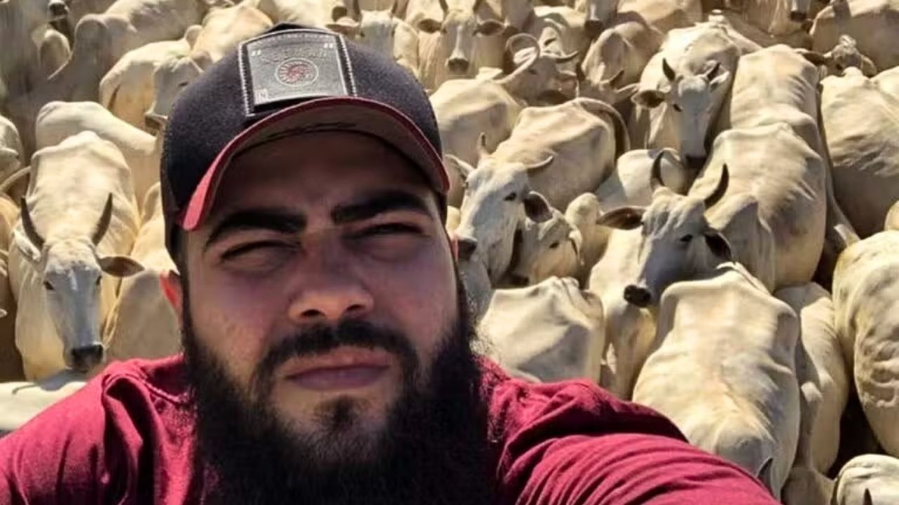 Cantor Henrique entre o rebanho Nelore da fazenda 'Terra Prometida', refúgio e império do agronegócio. Foto: Reprodução Homem barbudo de boné preto e camiseta vermelha tira selfie com gado Nelore branco ao fundo.