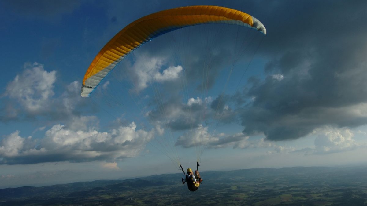 Pessoa em parapente amarelo e azul voando sobre vales verdes e montanhas sob c&eacute;u com nuvens.