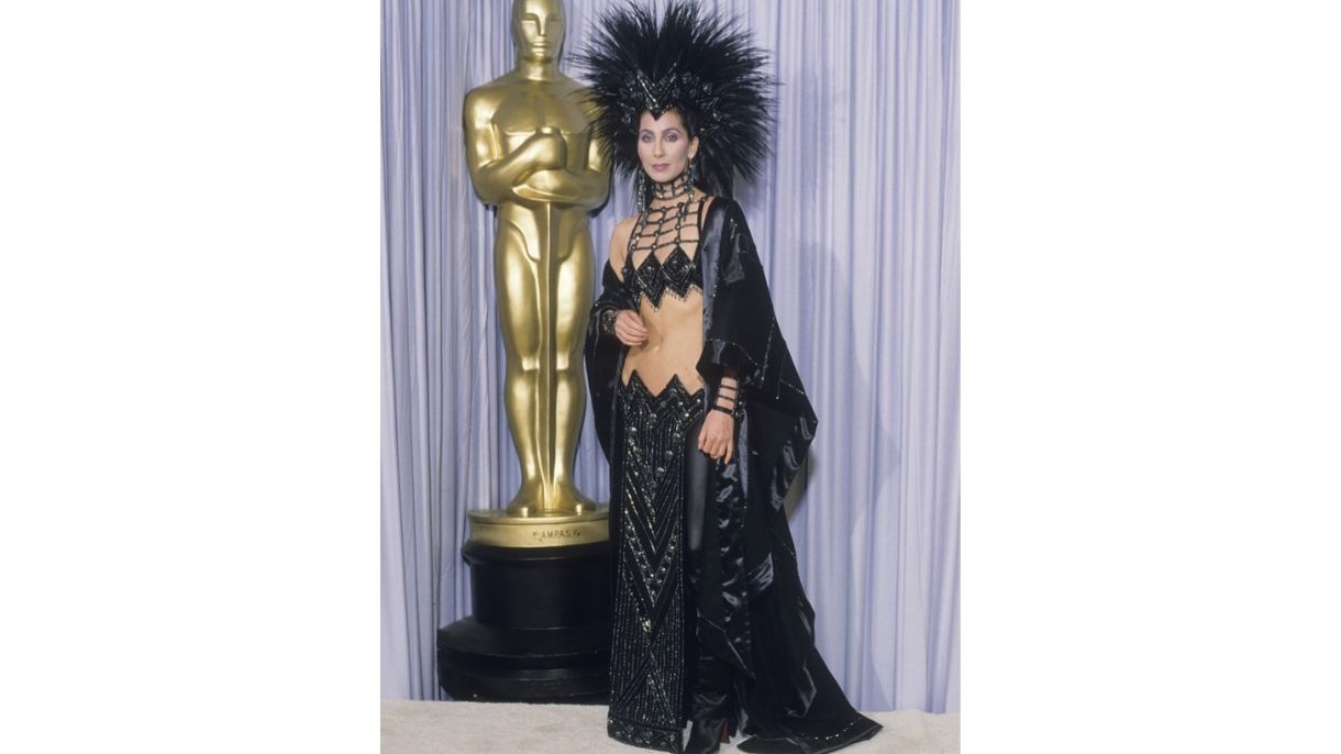 Cantora Cher em vestido preto extravagante e cocar de penas, posando com um Oscar.