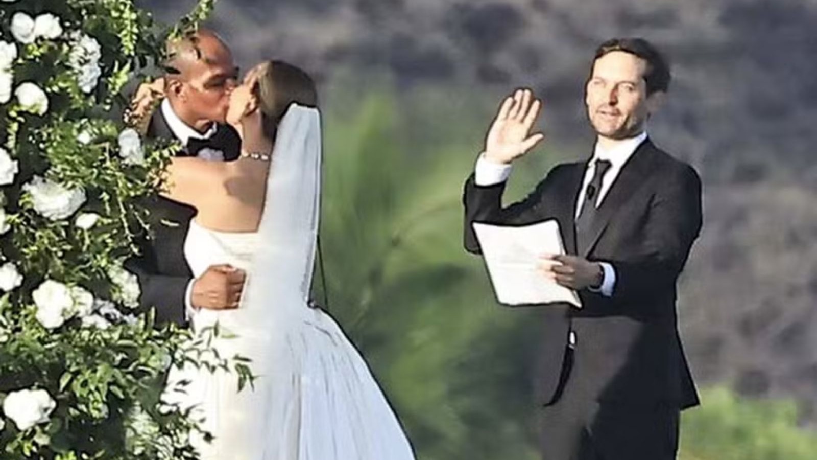 Casal se beijando no altar com Tobey Maguire &agrave; direita, erguendo a m&atilde;o, em casamento ao ar livre