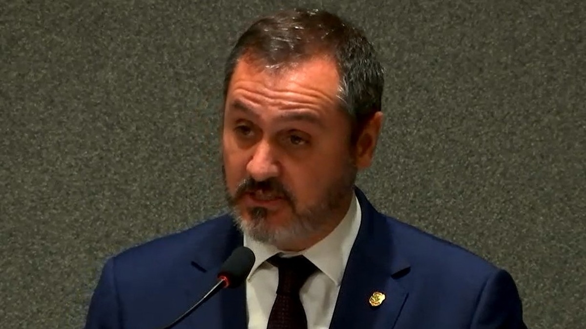 Foto: Reprodução Homem com barba e terno azul escuro, camisa branca e gravata, falando ao microfone