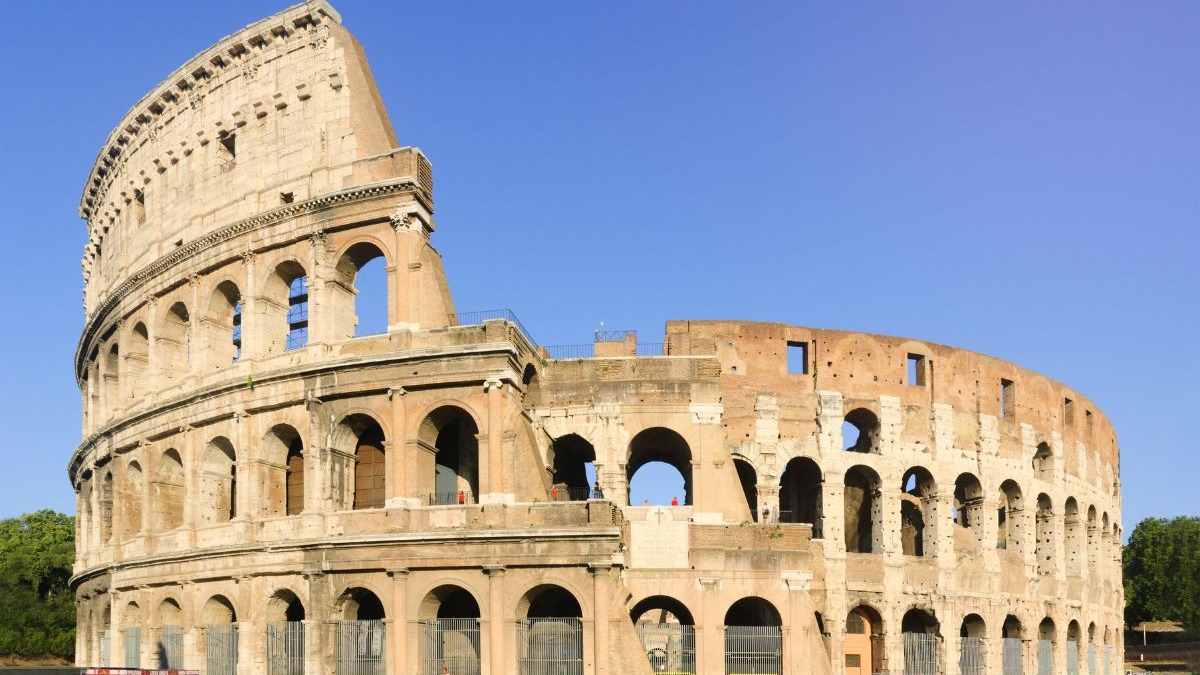 O Coliseu, maior anfiteatro do Império Romano, em Roma, figura como uma das 7 maravilhas do mundo moderno. Coliseu, anfiteatro de pedra com arcos e paredes parciais sob céu azul em Roma.