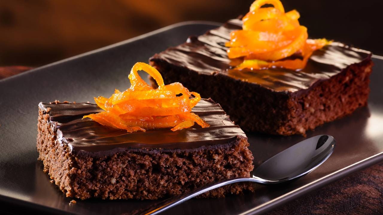 Prepare sobremesas como este brownie de chocolate com laranja para aproveitar o chocolate que sobrou da Páscoa. Créditos: depositphotos.com / colorvsbw Dois brownies de chocolate com cobertura e raspas de laranja caramelizada em um prato escuro.