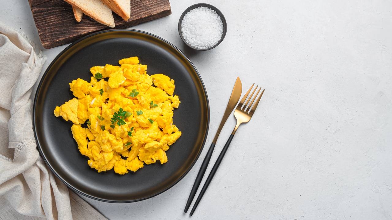 Ovos mexidos, um clássico proteico, são opção prática para a rotina de café da manhã sugerida para a quinta-feira. Ovos mexidos em prato preto, com torrada, saleiro e talheres dourados sobre mesa clara