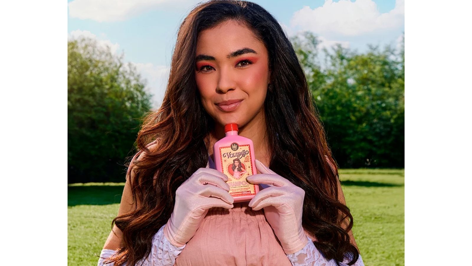 Mulher sorridente segura frasco rosa de xampu Lola Volum&atilde;o com pin-up em jardim ensolarado