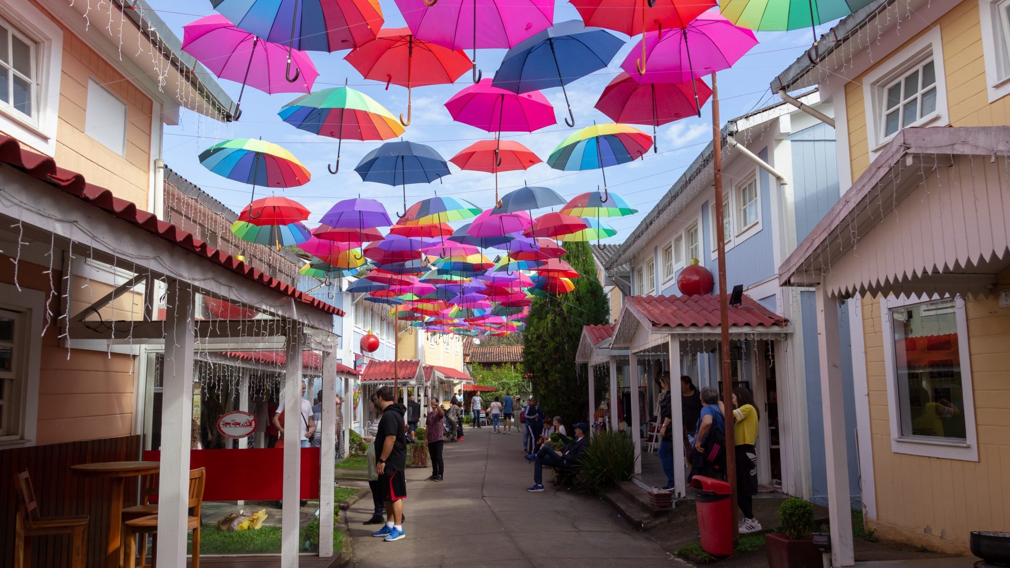 A decoração charmosa e a arquitetura inspirada na Europa são elementos-chave dos destinos brasileiros que encantam. Uma rua estreita com guarda-chuvas coloridos suspensos, edifícios coloridos e pessoas caminhando.