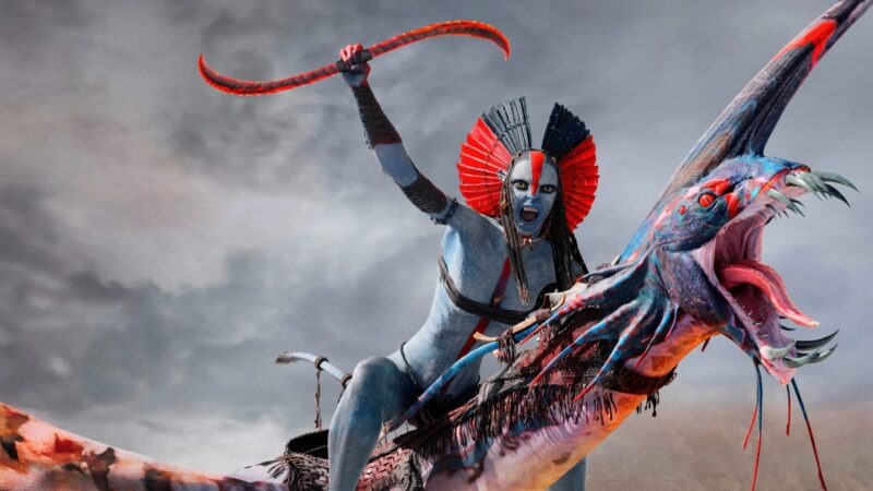 Na'vi azul com cocar vermelho, montado em criatura voadora azul e vermelha contra céu nublado.