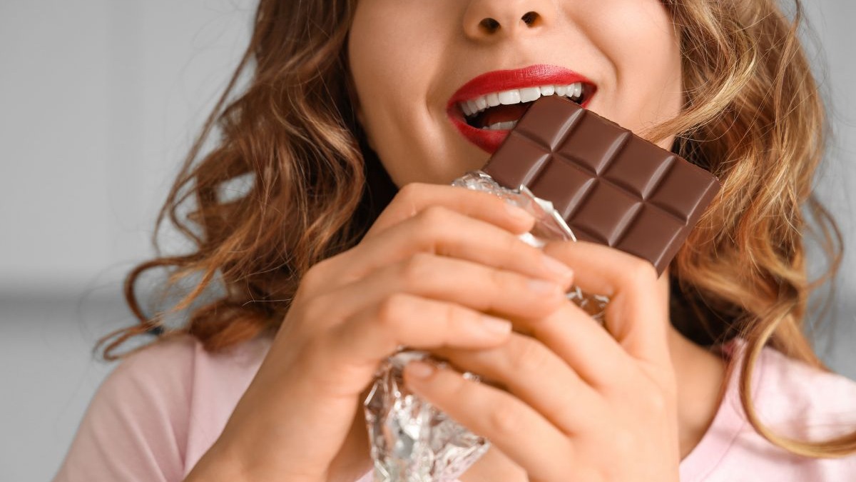 Close-up de mulher mordendo uma barra de chocolate com papel alumínio nas mãos.