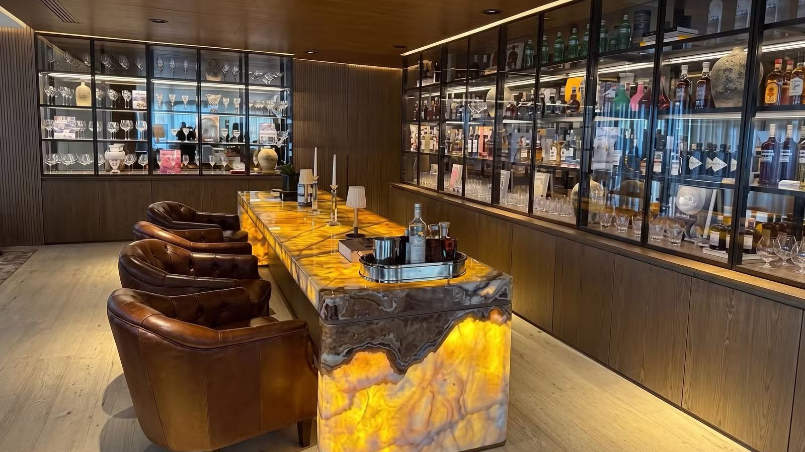 Bar de luxo com balcão de ônix iluminado, poltronas de couro e estantes com bebidas e taças