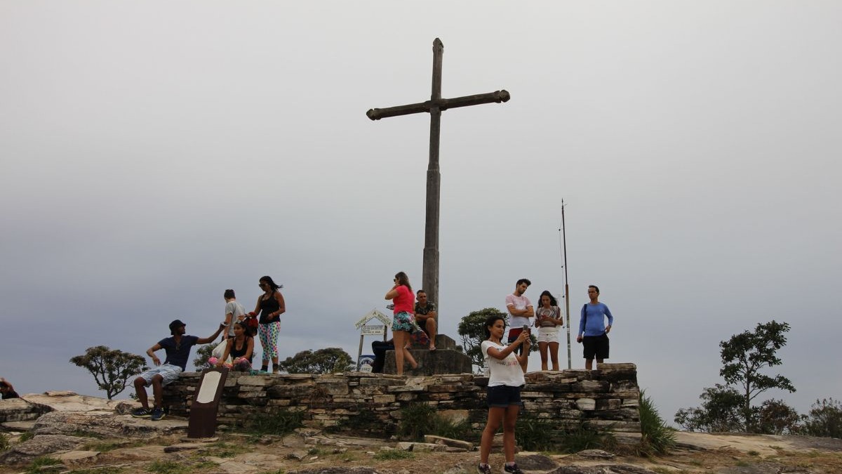 Pessoas em um mirante rochoso com uma grande cruz de madeira e c&eacute;u nublado, algumas tirando fotos.