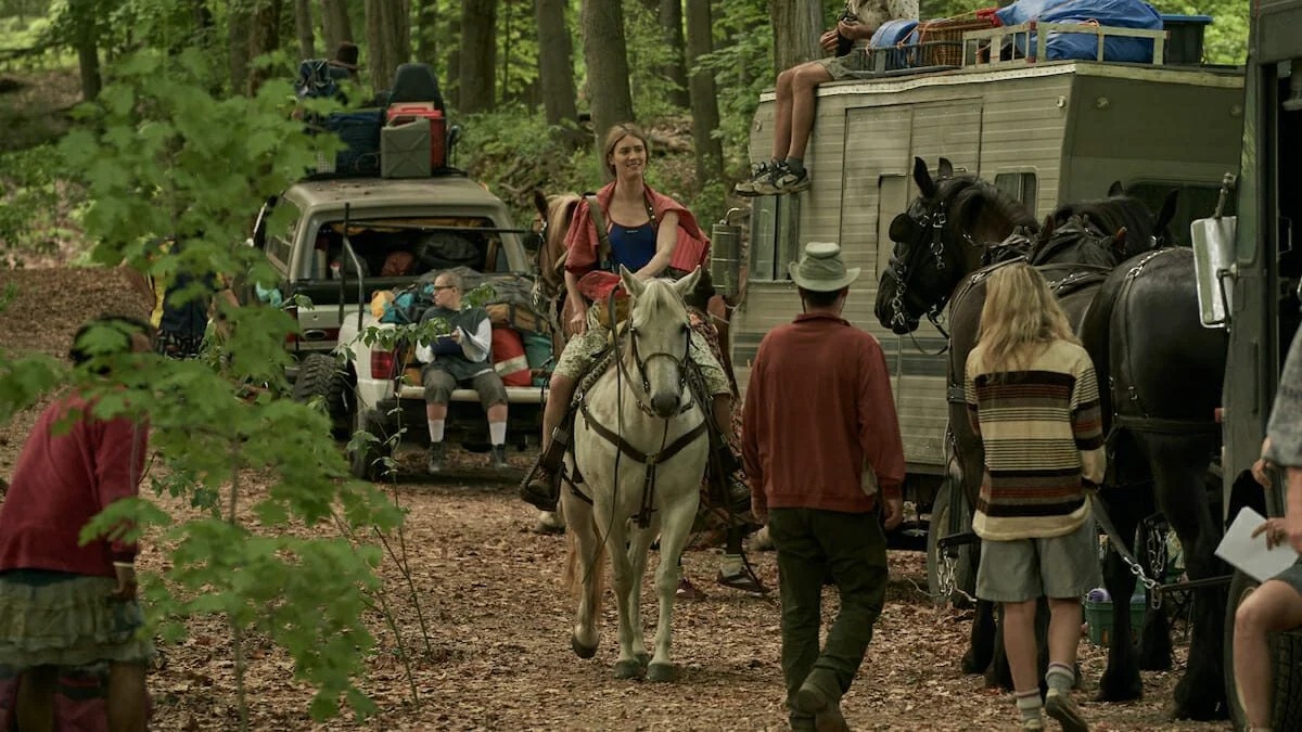 Mulher sorridente em cavalo branco lidera grupo com carro e trailer puxado por cavalos em floresta.