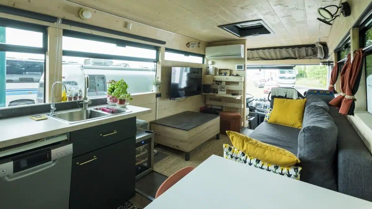 Interior de &ocirc;nibus motorhome com cozinha, sala de estar, sof&aacute; cinza, TV, lava-lou&ccedil;as e janelas