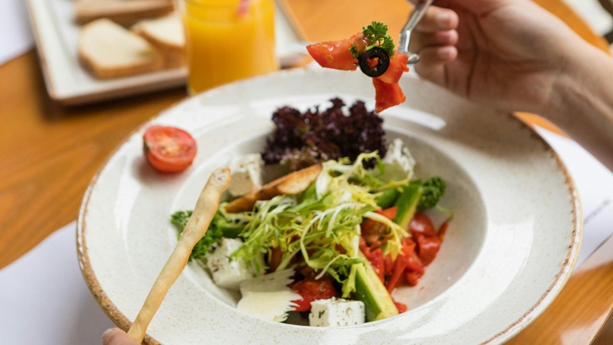 M&atilde;o com garfo e tomate acima de salada colorida com queijo, vegetais e p&atilde;o.