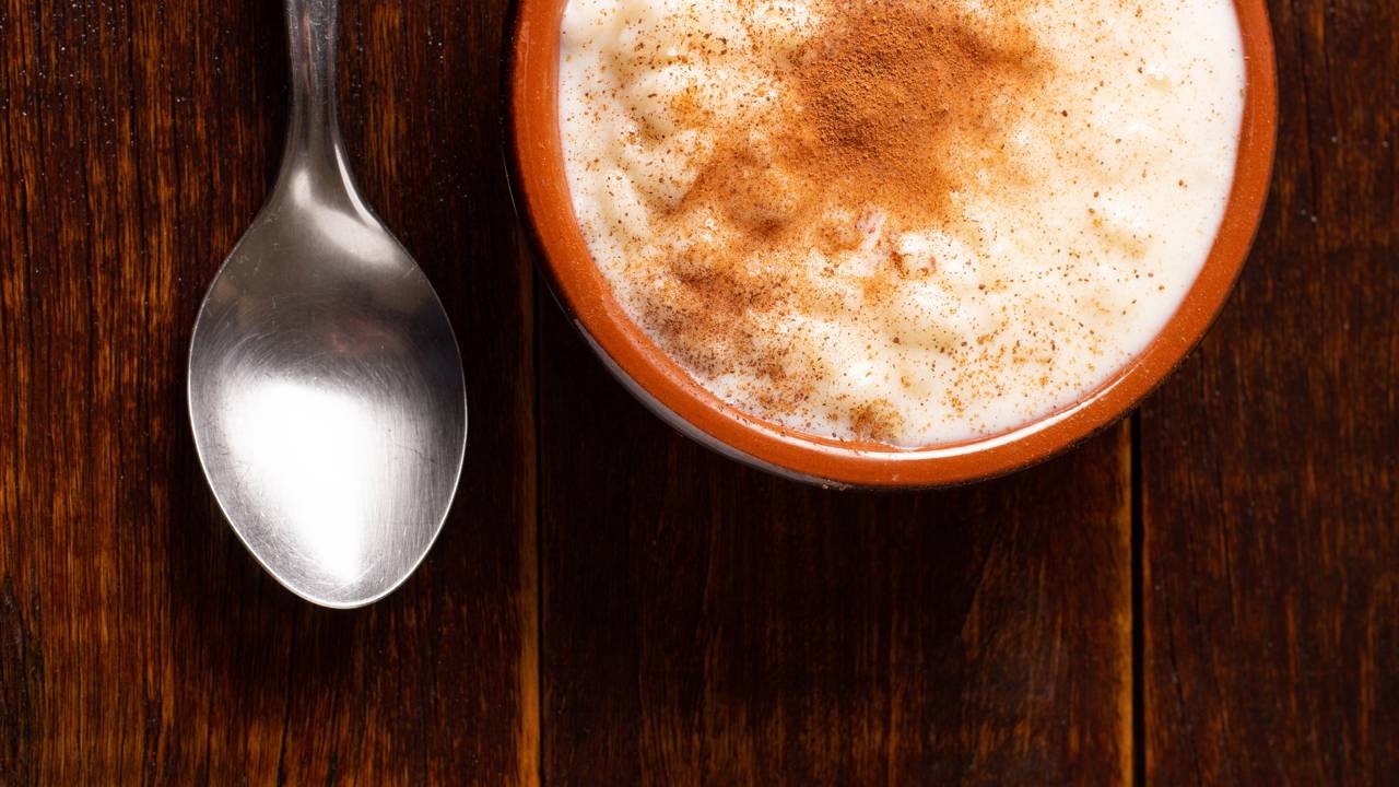 Aprenda a preparar a canjica cremosa, um doce tradicional perfeito para aquecer nos dias frios. Tigela de canjica cremosa com canela polvilhada e colher sobre mesa de madeira escura
