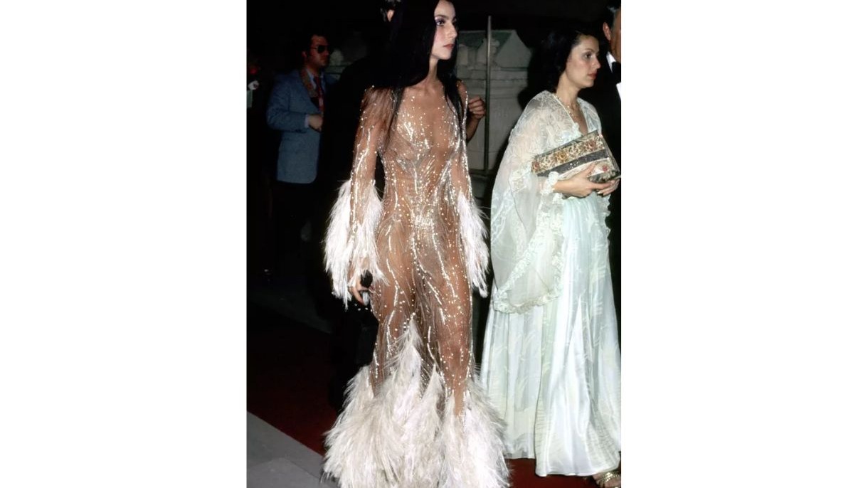 Cher usando um vestido transparente com brilhos e plumas brancas. Ao lado, uma mulher de vestido claro.
