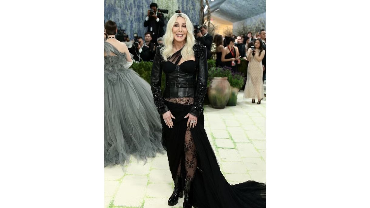 Cher sorri, usando jaqueta preta de couro, top corset e saia com fenda, em evento.
