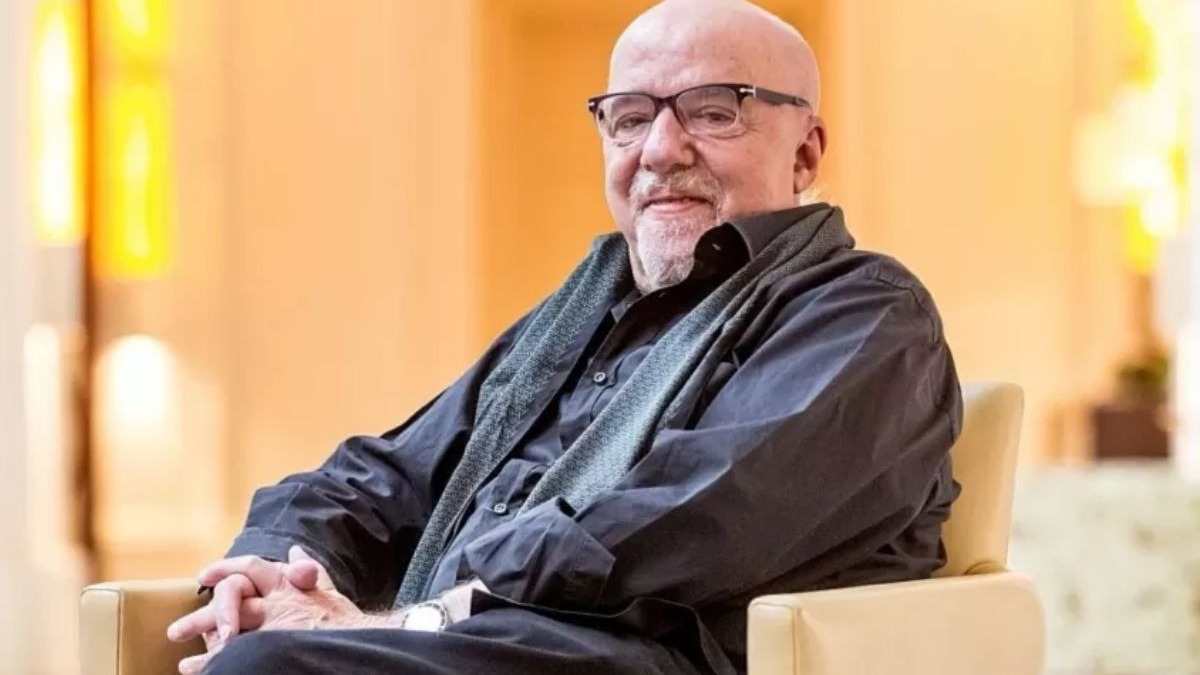 Homem calvo com óculos e barba, camisa escura, sentado em cadeira bege, sorrindo.