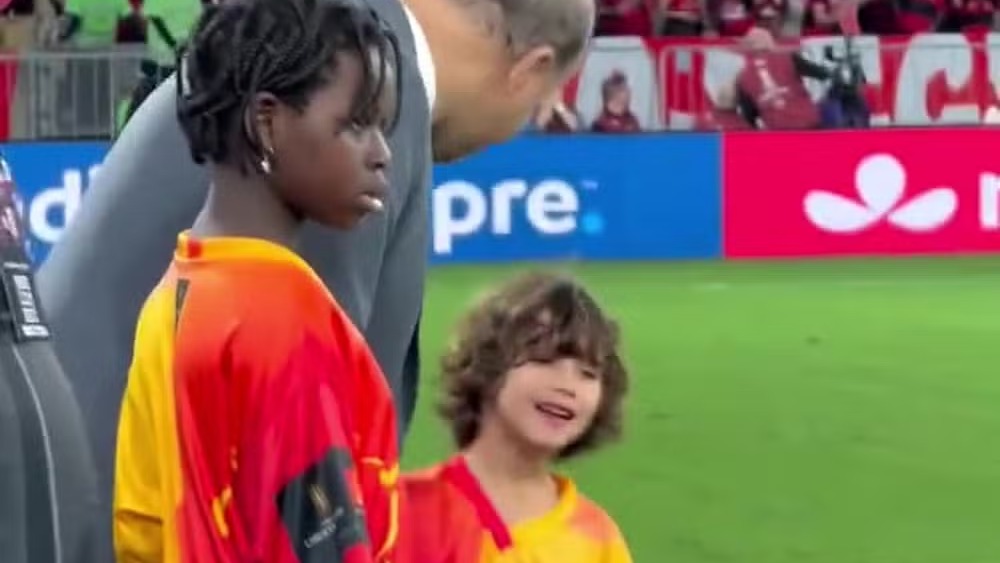 Bless e Zyan, filhos de Bruno Gagliasso, caminham em campo antes da partida do Flamengo na Libertadores. Dois meninos em campo, um de tranças, outro cacheado, vestindo camisas vermelhas e amarelas