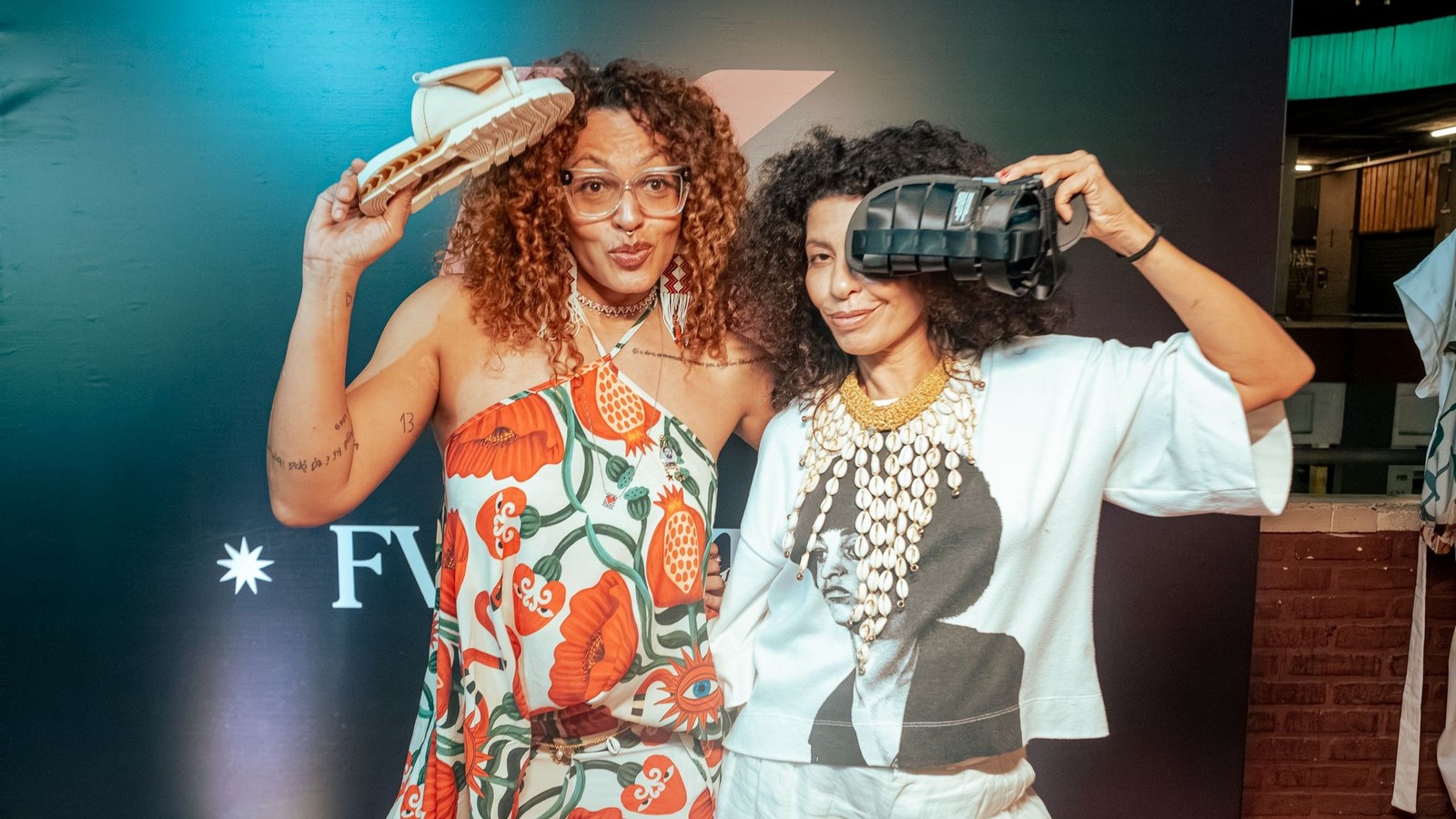 Duas mulheres de cabelo cacheado sorriem segurando sand&aacute;lias, uma branca e uma preta, em evento