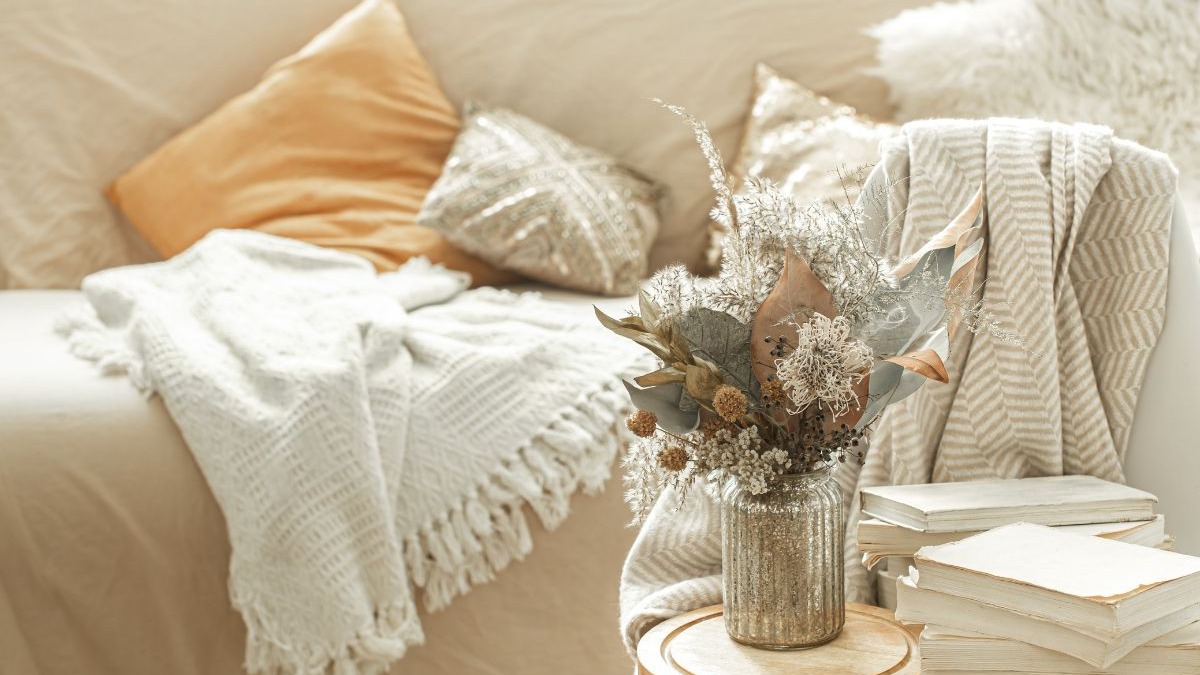Sofá com almofadas, manta, vaso de flores secas e livros em uma mesa lateral.