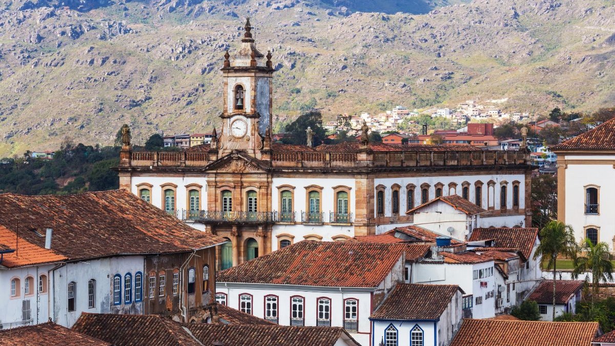 Vista a&eacute;rea de cidade colonial com edif&iacute;cio barroco e torre do rel&oacute;gio, telhados de barro, montanhas.