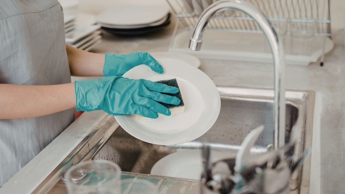Mãos com luvas turquesa lavando um prato branco com esponja em pia de inox.