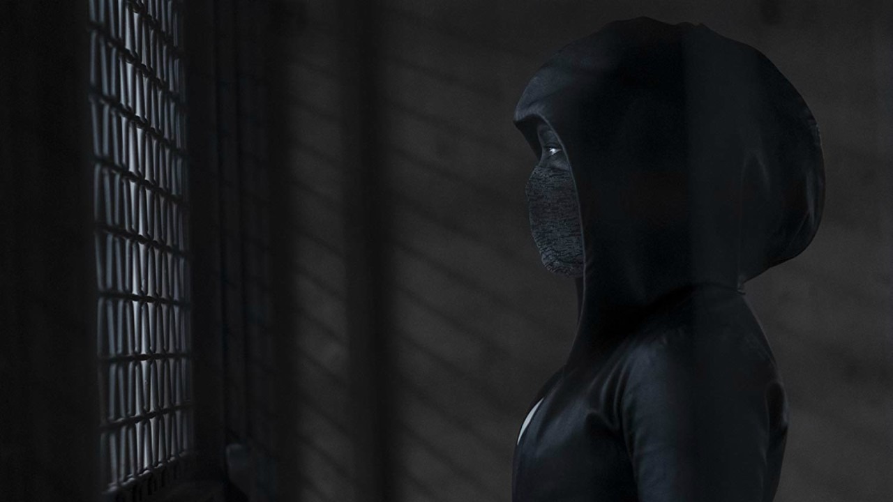 A complexa série Watchmen, recomendada para fãs de 'The Boys', explora vigilantes mascarados e a moralidade em um mundo falho. Figura encapuzada e mascarada com um olho visível, olhando por uma grade escura.