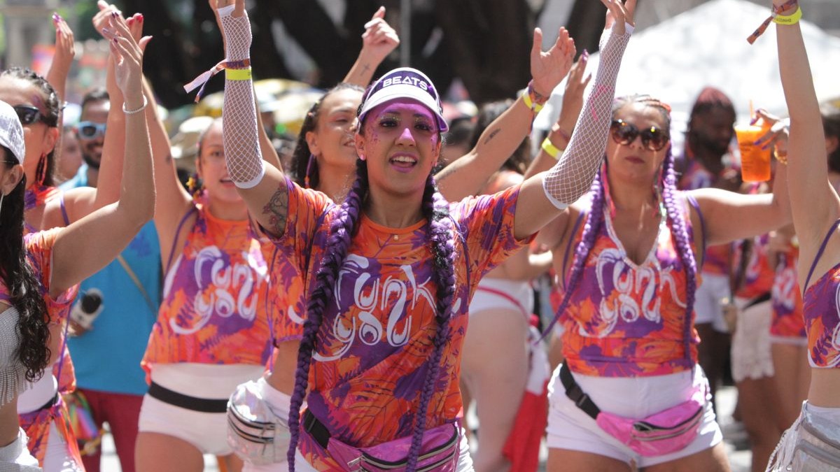 Mulher sorrindo com tranças roxas e camisa 'Funk', braços erguidos em festa de carnaval.