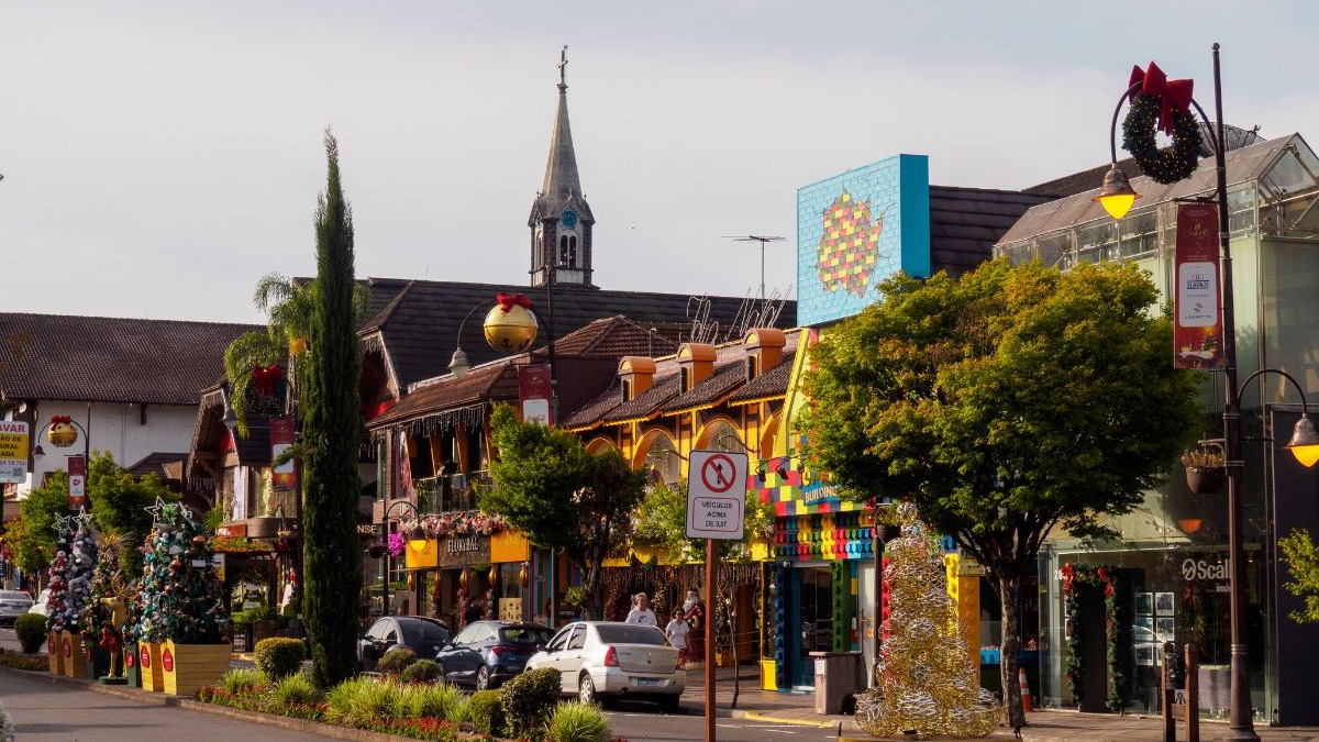 Rua de cidade com arquitetura europeia, &aacute;rvores, carros, decora&ccedil;&otilde;es natalinas e um campan&aacute;rio ao fundo.