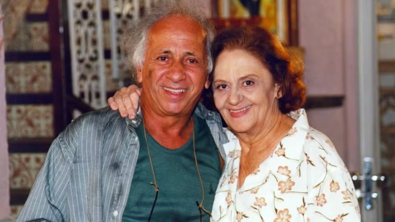 Homem e mulher idosos sorrindo para c&acirc;mera, ele de camisa listrada, ela de blusa florida