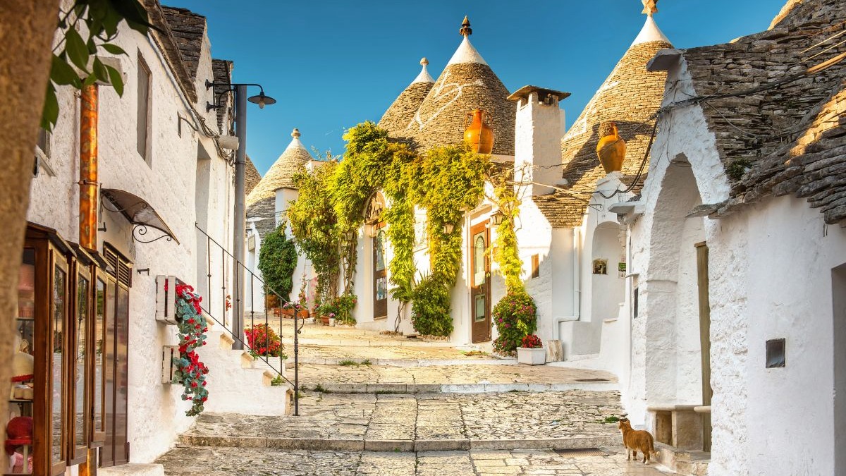 As charmosas casas trulli de Alberobello, na Puglia, são um Patrimônio Mundial da UNESCO e um destaque do turismo na Itália. Rua de casas brancas com telhados cônicos de pedra, vegetação e um gato, em Alberobello.
