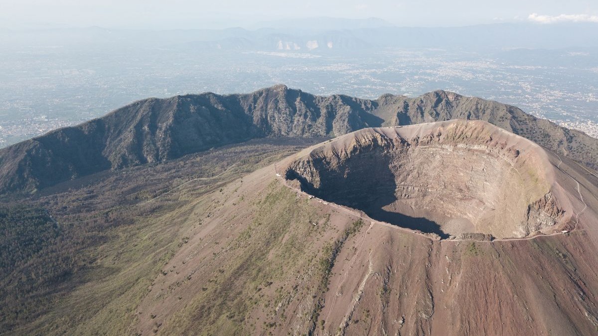 A trilha para a borda da cratera do Vesúvio, na Itália, permite aos visitantes admirar a vista da baía de Nápoles, um dos destaques do parque nacional. Cratera do Vesúvio com trilha na borda, montanhas e cidade distante sob céu claro.