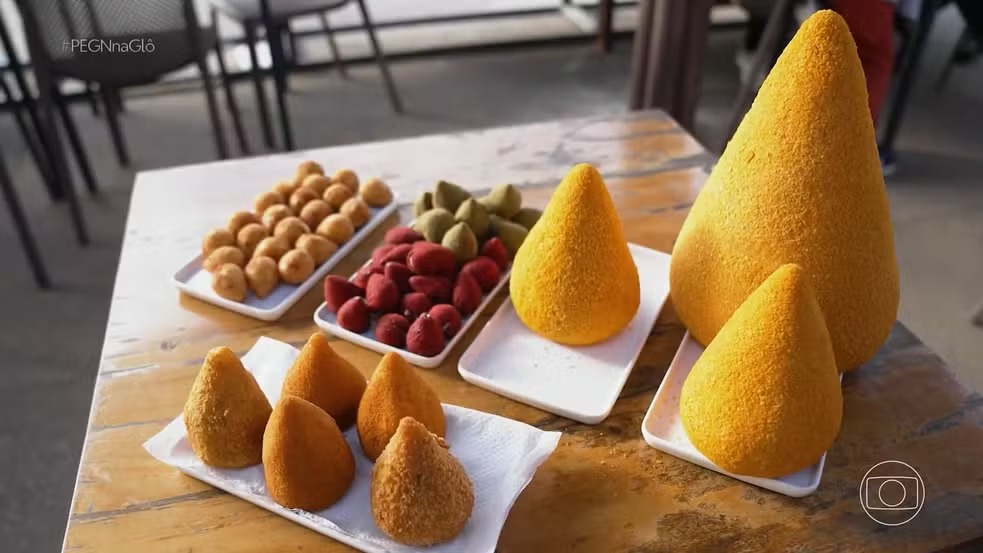 A variedade é destaque no negócio de Marlene, que produz coxinhas de diversos tamanhos e sabores, incluindo versões gigantes. Coxinhas variadas em pratos brancos, com morangos e doces verdes, em mesa de madeira