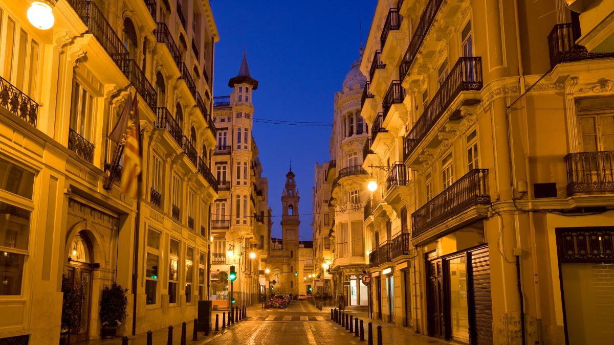 Rua estreita de Val&ecirc;ncia &agrave; noite, edif&iacute;cios iluminados, bandeira e torre sineira ao fundo.