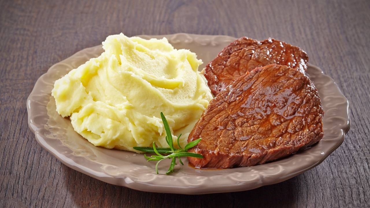 O purê de batata light é uma das receitas leves para aquecer o inverno. Prato com purê de batata, dois pedaços de carne e ramo de alecrim sobre mesa de madeira