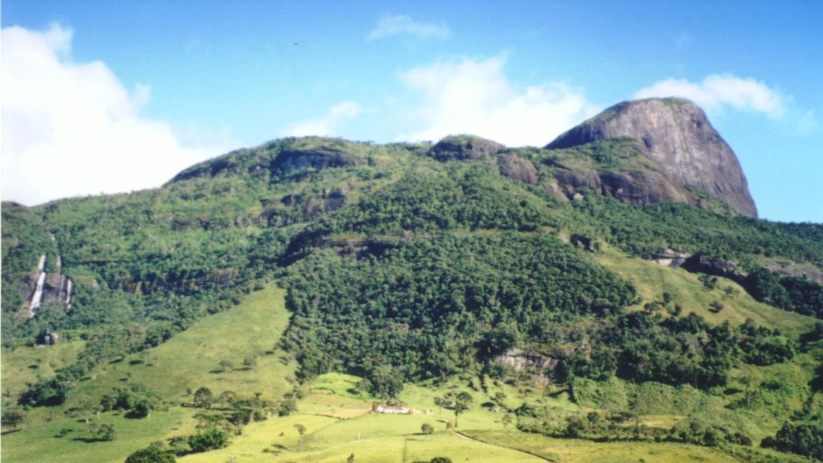 Montanha verde exuberante, pico rochoso, cachoeiras e vale com campos verdes e casas.