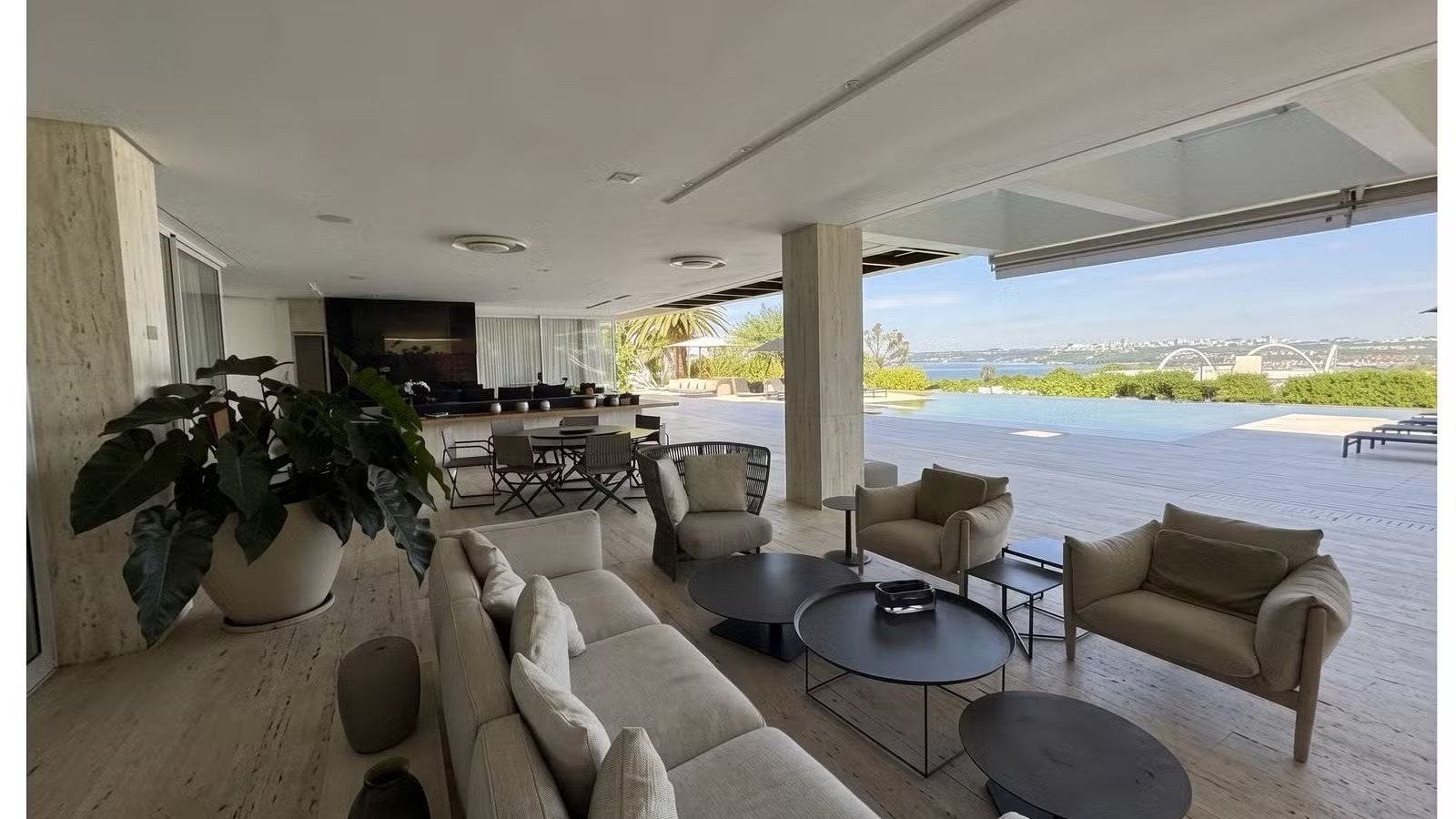 Ampla sala de estar com sofá, poltronas, mesas, planta, piscina e vista para lago, ponte