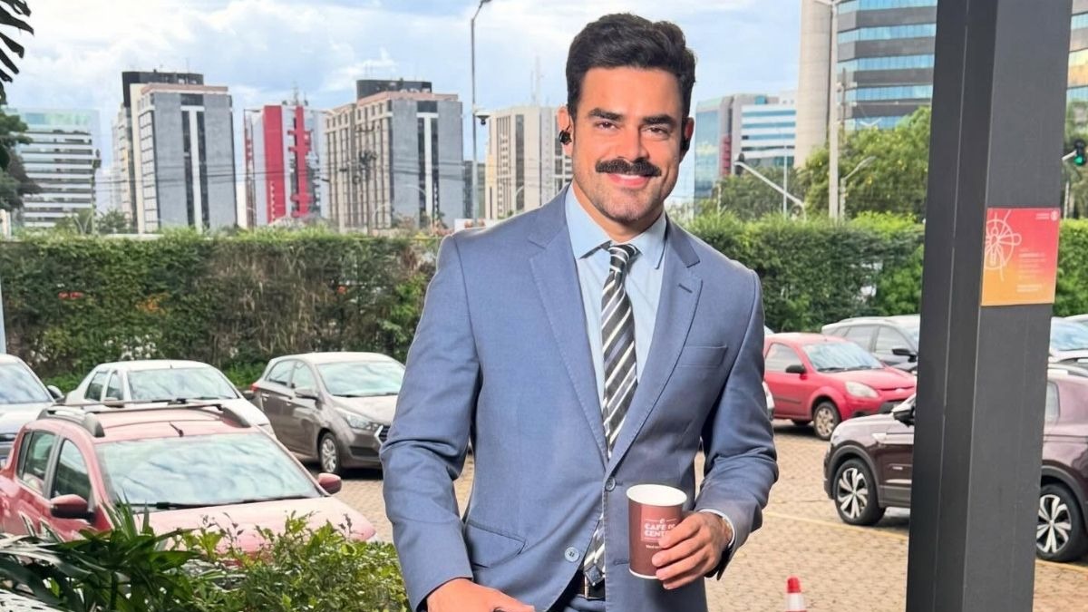 Túlio Amâncio, novo comentarista político do SBT News, após sua saída da Globo e passagem pela GloboNews. Foto: Reprodução Jornalista Túlio Amâncio de terno azul, gravata listrada, bigode e fones, sorri, segurando copo de café