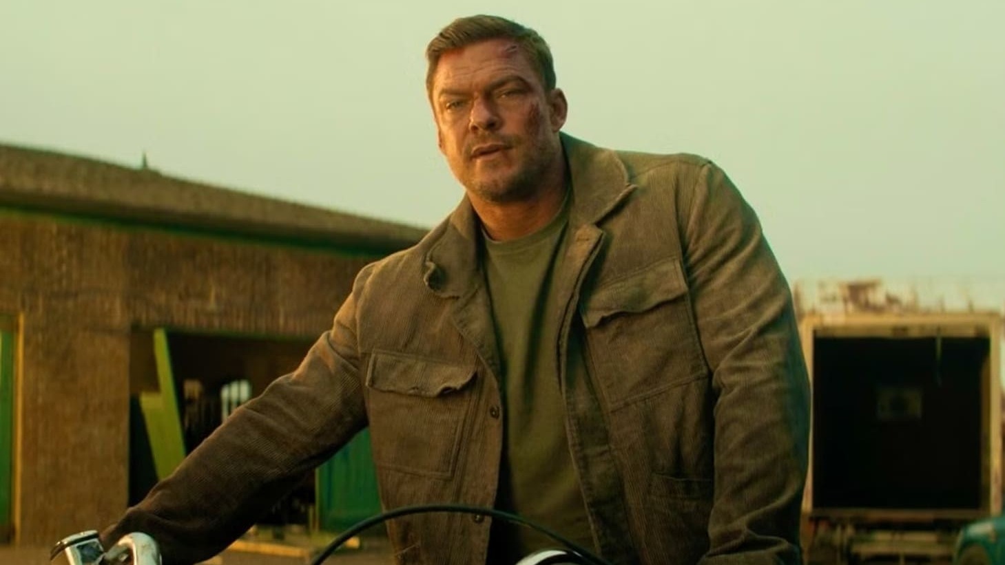Alan Ritchson interpreta o protagonista Jack Reacher, que retorna para a aguardada quarta temporada da série de sucesso. Alan Ritchson com jaqueta marrom, rosto arranhado, em moto contra fundo industrial.