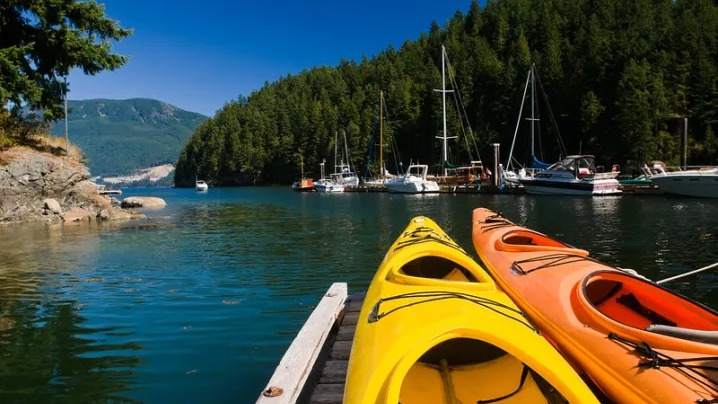 Em locais como este, repletos de natureza e atividades aquáticas, são gravadas as cenas idílicas de 'Virgin River' no Canadá. Caiaques amarelo e laranja num cais de madeira, com barcos, floresta e montanhas ao fundo.